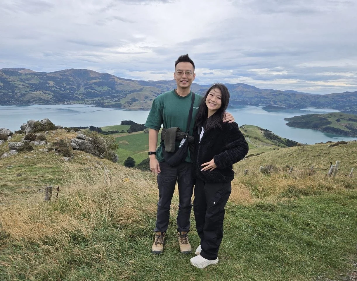 Two visitors standing on a hillside above Akaroa Harbour after visiting Pōhatu